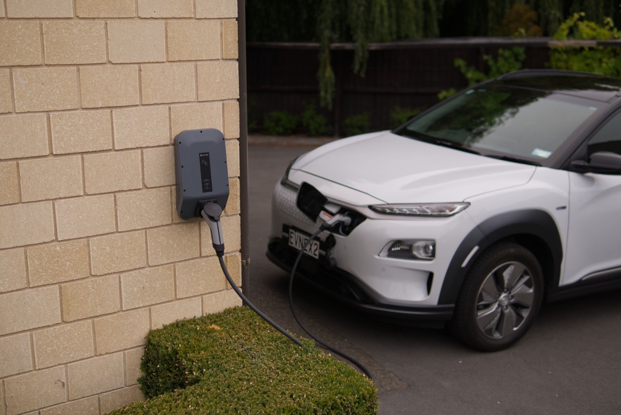 An electric car charging from a wall-mounted home charger on a house drive